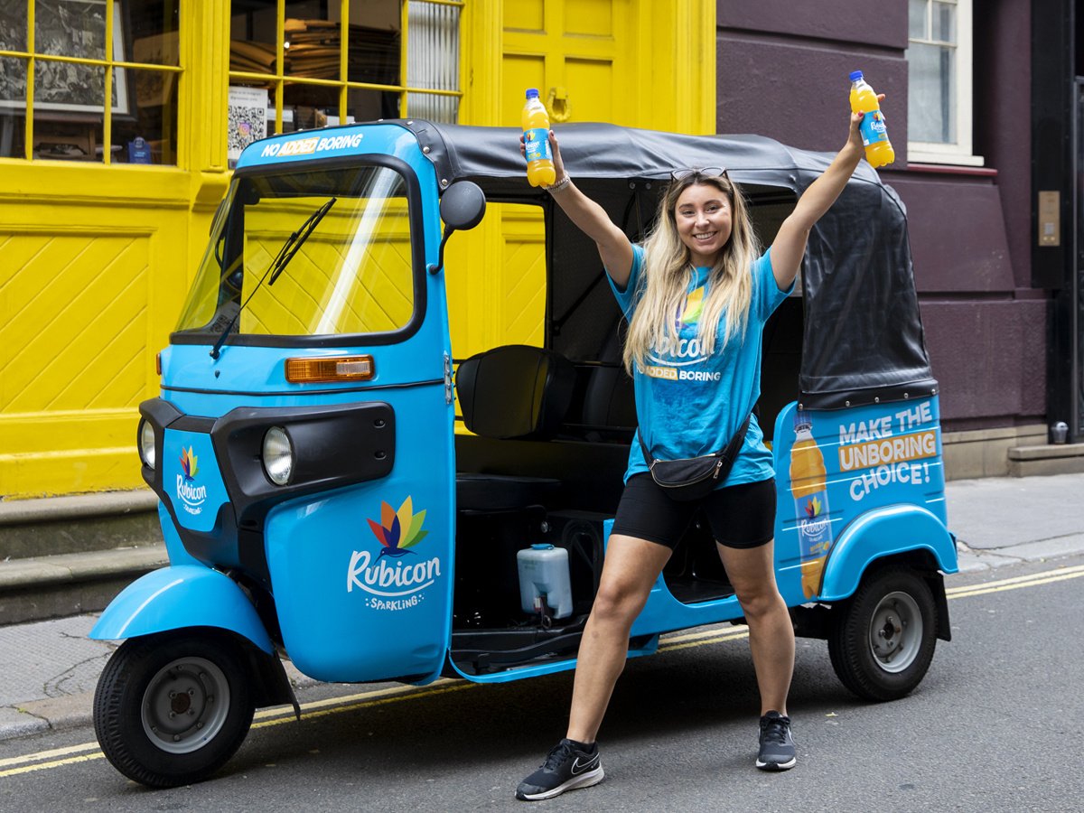 A staff member holds drink samples in front of the blue Rubicon promotional tuk tuk hire