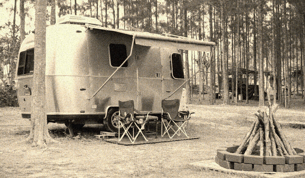 A grainy image of a silver Airstream caravan in the forest with deckchairs and a fireplace.