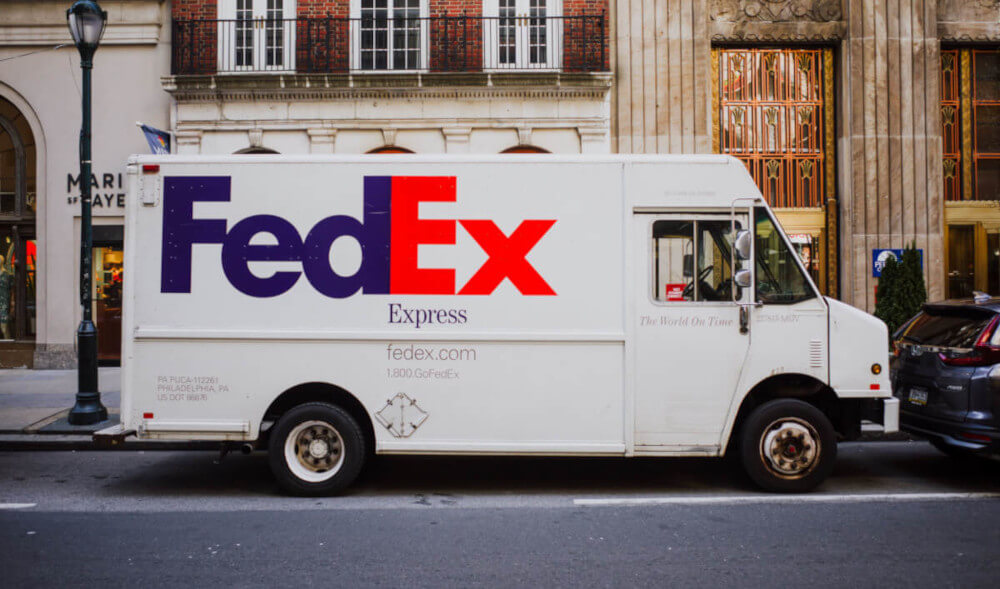A white FedEx truck on a city street, with branding on the side of the delivery vehicle. Now available for hire.