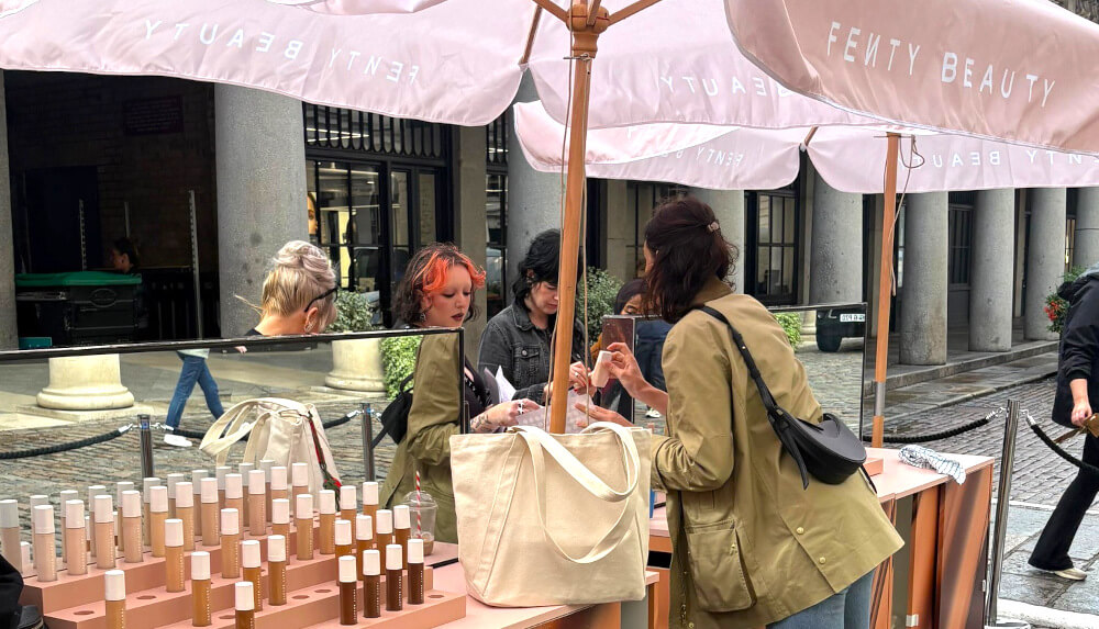 Branded parasols and cosmetic displays for Fenty, with staff helping a guest.