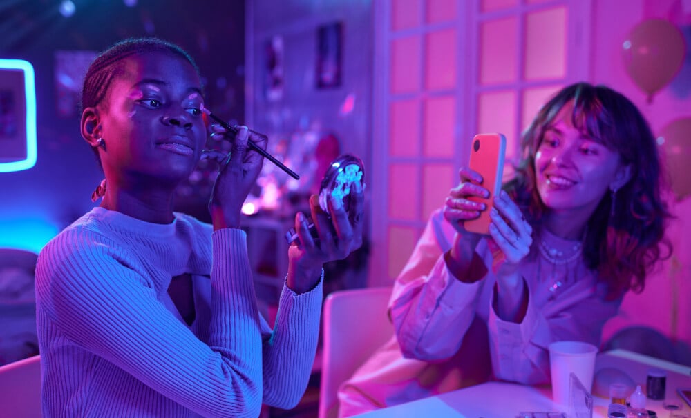 An influencer puts on makeup for a vlog while another films on her phone.