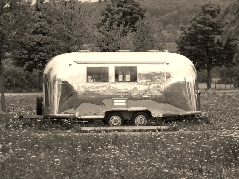 A sepia image of a silver vintage Airstream trailer in a field, as invented by Wally Byam.